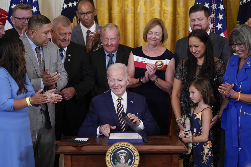 President Biden sits at desk smiling and holding PACT Act bill he just signed as he is surrounded by smiling advocates, veterans and their families looking down at him.